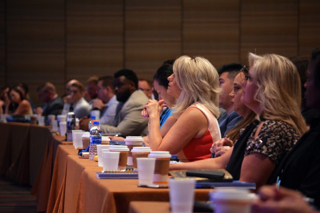 attendees of conference sitting at tables with coffee and waters all facing towards the presenter